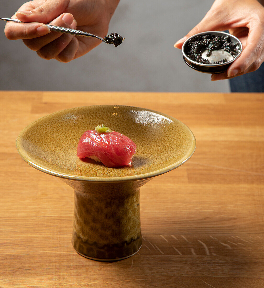 A close-up image showing hands using tweezers to garnish a piece of sushi with caviar, placed delicately on a textured ceramic plate