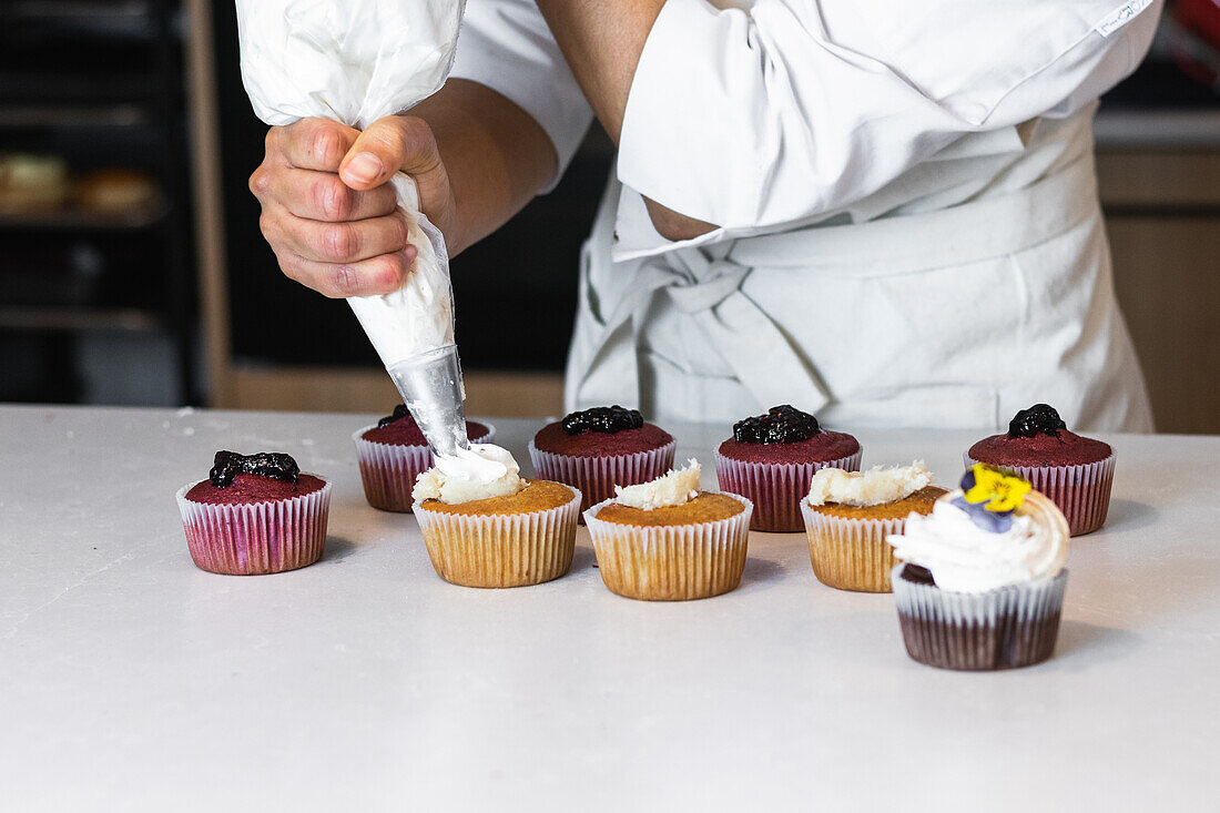 Unrecognizable crop baker squeezing whipped cream from pastry bag on tasty cupcakes while cooking desserts in bakehouse