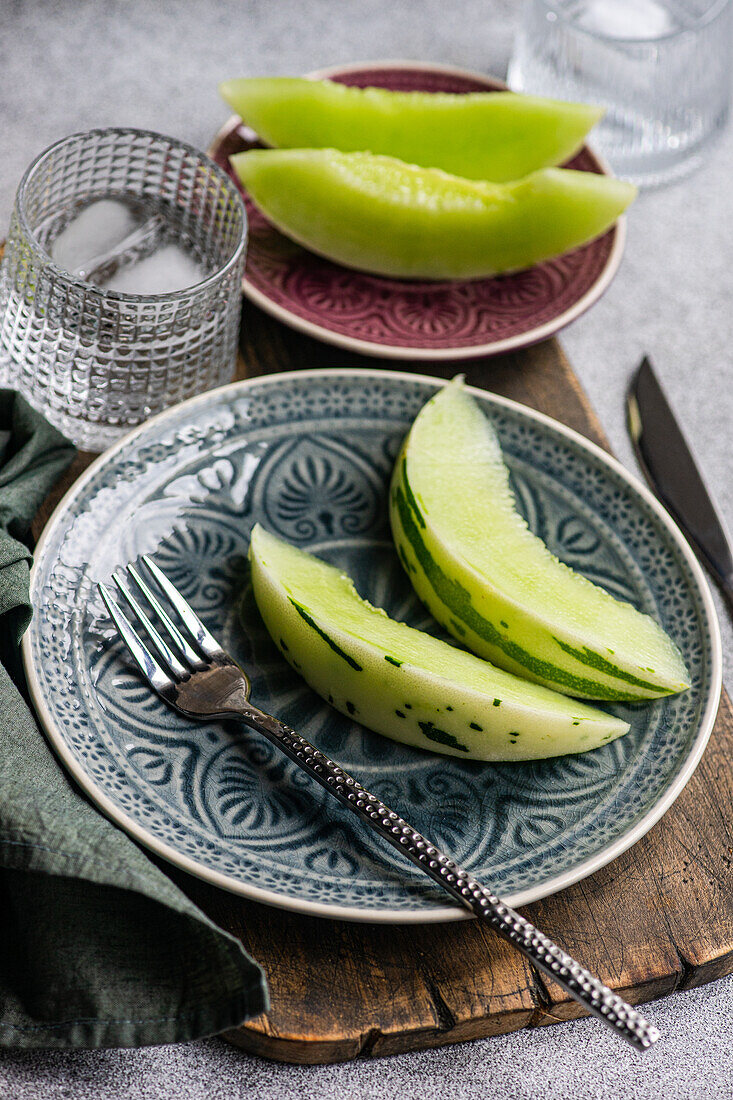 From above organic mini melon slices arranged on ornate ceramic plates, accompanied by a glass of water with ice and a fork on a textured tablecloth.