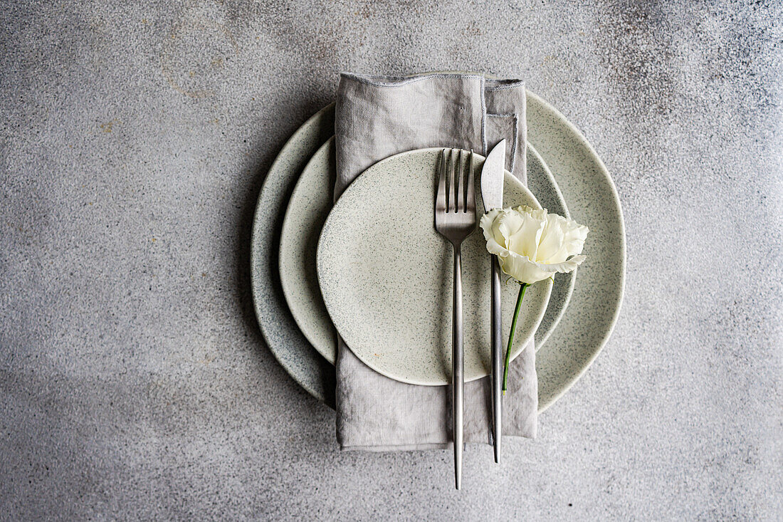 Top view of tasteful table setting featuring a stack of neutral colored plates, a neatly folded gray linen napkin, and stainless steel cutlery, adorned with a delicate white Godetia flower, presented on a textured gray background