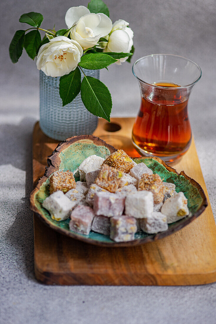 An assortment of colorful Traditional Turkish delights on a plate with a glass of black tea, complemented by a vase of fresh white roses