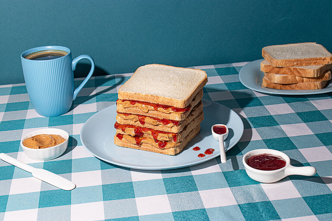 Ein Stapel frisch zubereiteter Erdnussbutter-Gelee-Sandwiches mit einer Tasse amerikanischem Kaffee und einigen Brotscheiben vor einem blauen Hintergrund