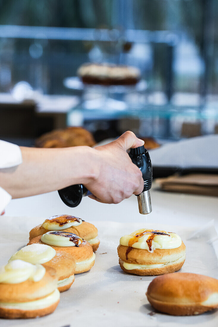 Unrecognizable crop chef caramelizing sugar cream on sweet Berliners with gas torch while preparing vegan dessert in bakehouse