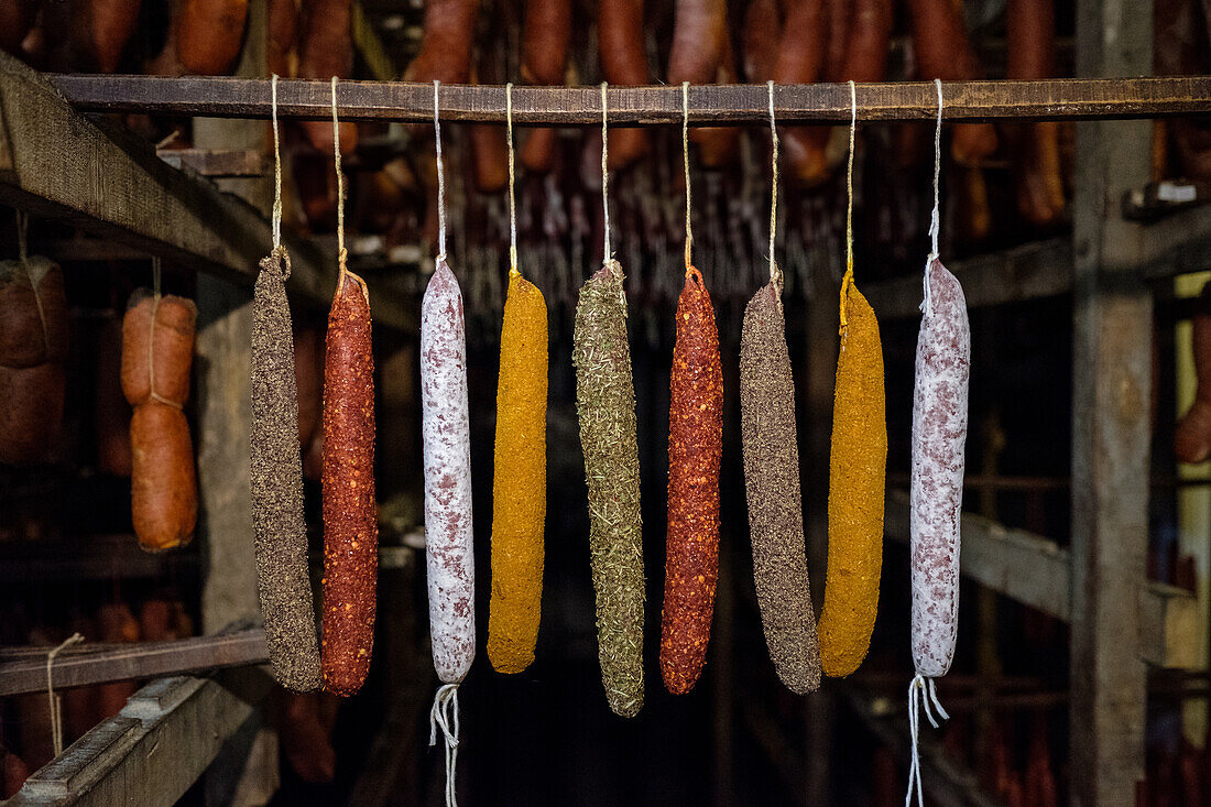 A selection of cured sausages, including sobrasada, hangs in a rustic smokehouse. The sausages are seasoned with various spices, showcasing a range of colors from deep red to yellow.