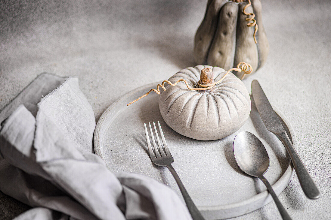 Ein geschmackvoll arrangiertes, herbstliches Flatlay mit einem grauen Essteller, einer Leinenserviette, Kürbisdekorationen und Silberbesteck auf einer strukturierten Oberfläche