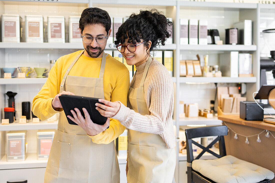 Two multiethnic employees in aprons are engaged with a digital tablet amidst a brightly-lit shop.