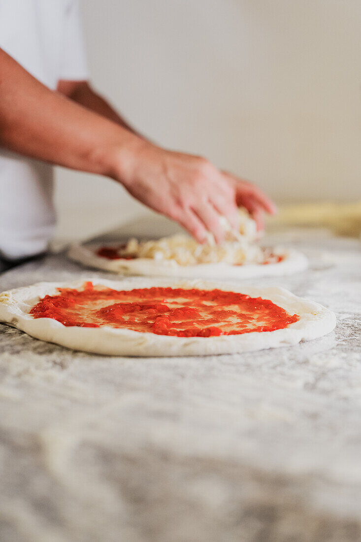 Ein anonymer Koch bereitet in einer Restaurantküche eine Pizza zu, indem er Soße und Käse auf den Teig streicht, und zeigt einen gesichtslosen und konzentrierten kulinarischen Moment