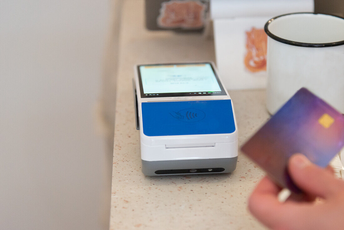 A cropped unrecognizable view of a hand holding a credit card near a modern payment terminal displaying a contactless payment interface.