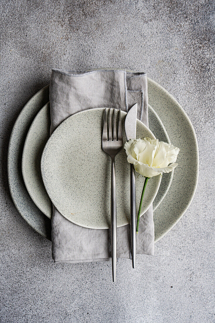 Top view of elegant table setting featuring speckled ceramic plates stacked with a linen napkin and sleek cutlery, complemented by a delicate Godetia flower, all arranged on a textured gray background