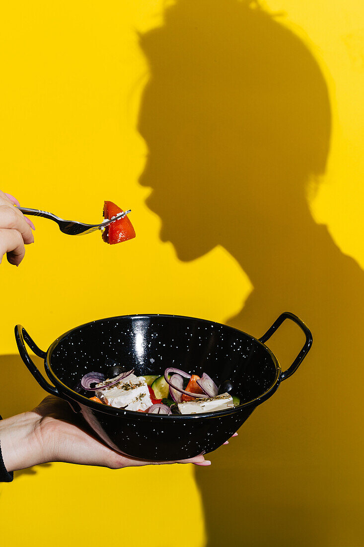 Eine Hand hält einen schwarzen Wok, der mit bunten vegetarischen Beilagen wie Tomaten und Zwiebeln gefüllt ist und sich von einem leuchtend gelben Hintergrund abhebt, der gesunde Grilloptionen hervorhebt