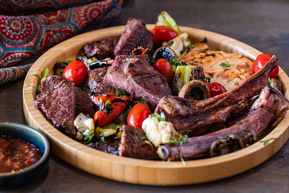 A generous wooden platter loaded with perfectly grilled meats including ribs and lamb chops, paired with colorful grilled vegetables like tomatoes and peppers