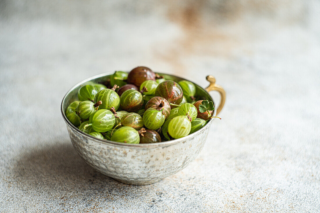 From above, a variety of fresh organic gooseberries in hues of green and burgundy rest in an ornate metal bowl on a textured grey surface, with subtle lighting enhancing their natural sheen.