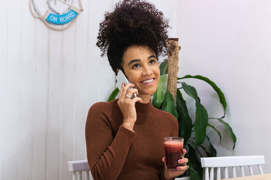 Happy African American female freelancer speaking on smartphone and laughing while sitting at table with smoothie in cafe and working on remote project