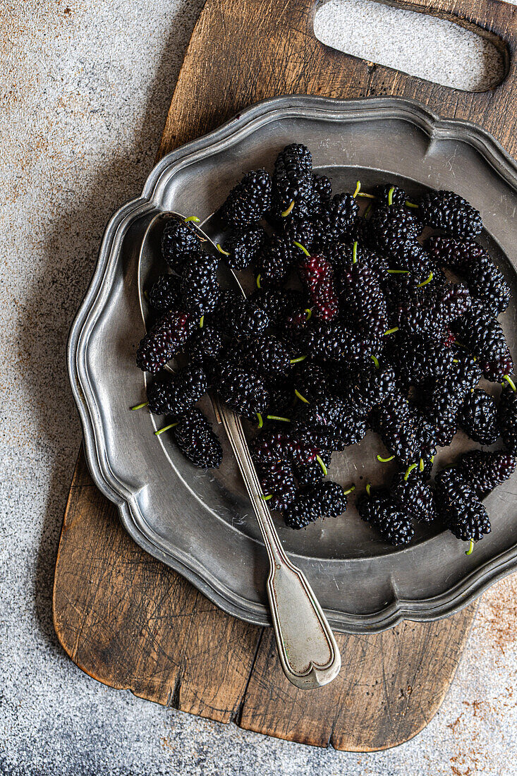 Top view of a rustic setting showcases fresh juicy mulberries in a vintage silver tray, resting on a weathered wooden chopping board, complemented by a textured silver spoon and a dusty backdrop.