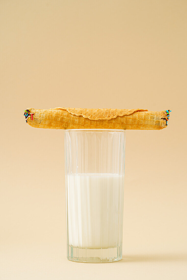 A crispy waffle roll filled with condensed milk resting atop a full glass of milk, set against a pastel background.