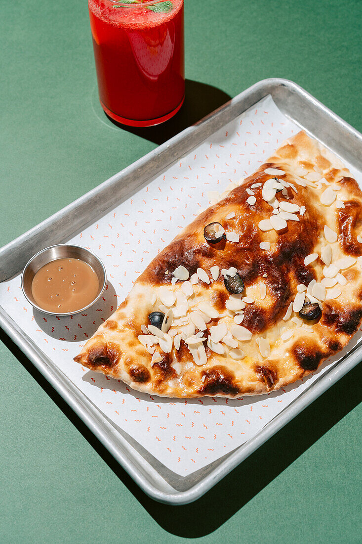 Ein süßer gebackener Cheburek garniert mit Mandelblättchen, gepaart mit einem erfrischenden Glas Beerenlimonade, präsentiert auf einem Metalltablett.