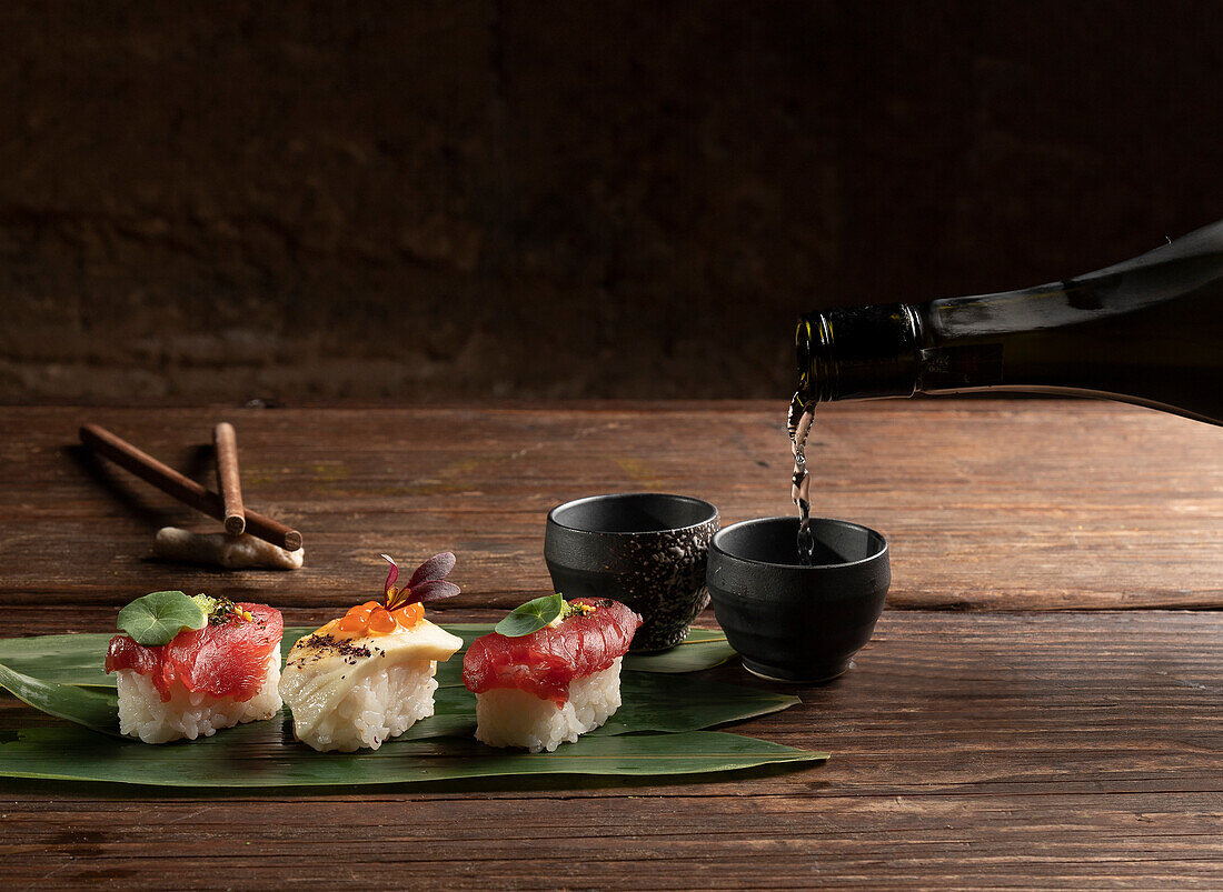 A sophisticated display of sushi on a leaf with sake being poured into a small cup, highlighting a traditional Japanese culinary scene on a rustic wooden table