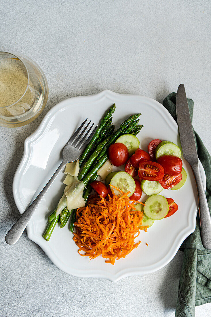 From above, a nutritious plate of asparagus spears, sliced tomatoes, cucumber rounds, and grated carrot topped with slices of cheese. A white plate serves this vibrant mix, with utensils on the side, offering a balanced meal option.