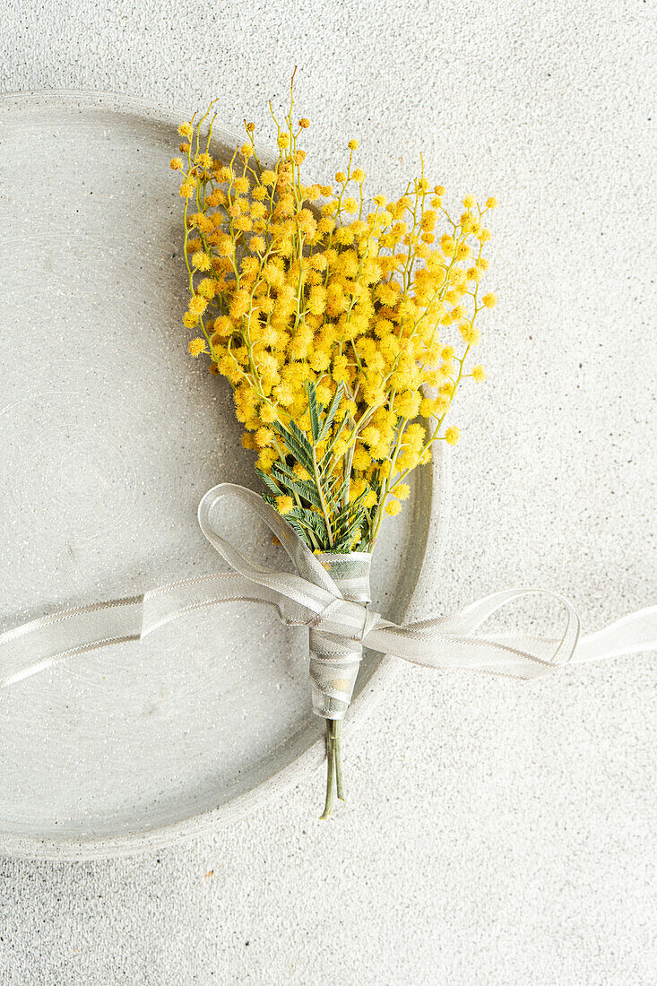 Top view of delicate arrangement of vibrant mimosa flowers tied with a silver ribbon, presented on a sleek, textured table surface for a sophisticated touch