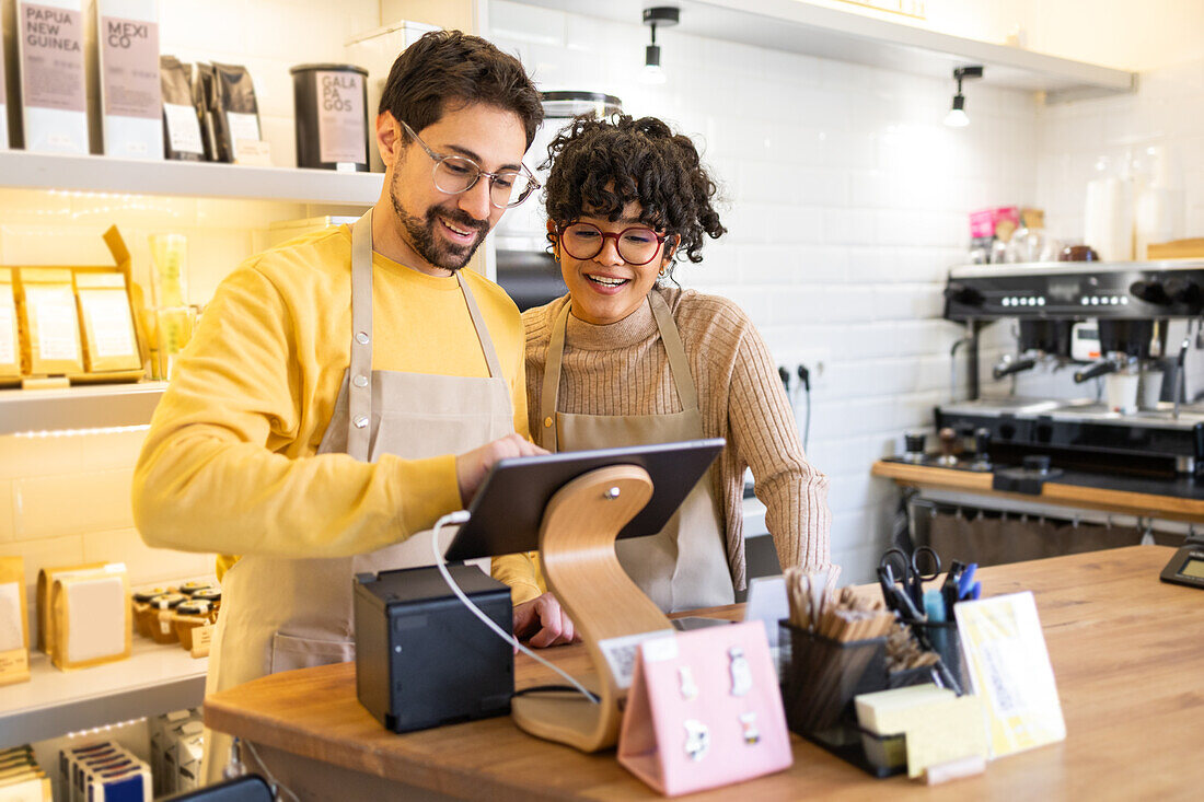 Smiling male and female multiethnic baristas collaborate using a tablet in a cozy, well-lit café setting, displaying teamwork and technology in small business.