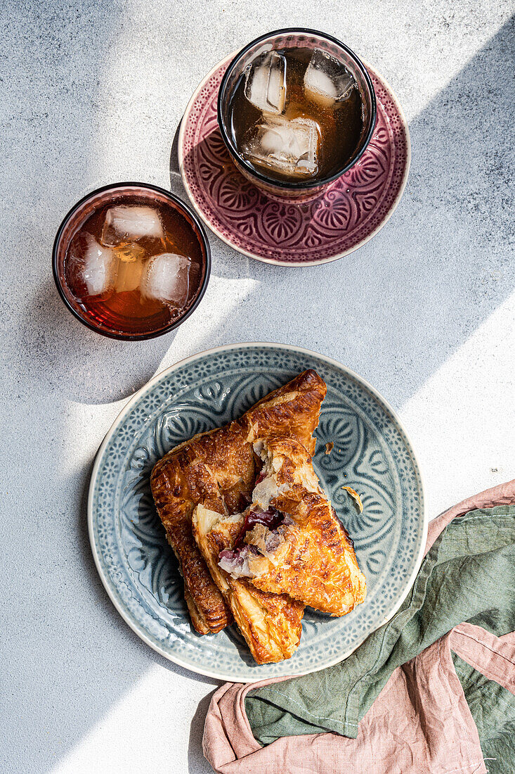 Top view of two glasses of ice tea and a slice of cherry pie served on a decorative ceramic plate, illuminated by natural sunlight.