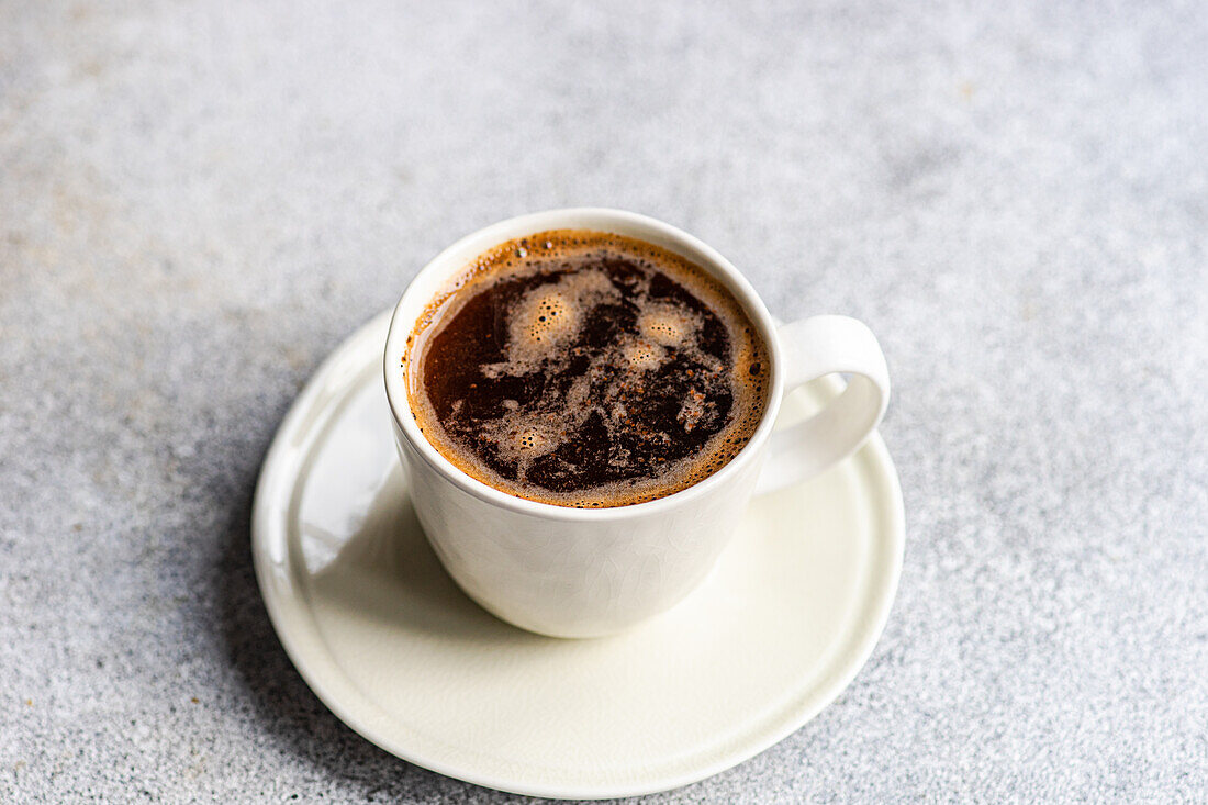 A From above of a freshly brewed cup of black coffee, with visible bubbles, served in a white ceramic cup seated on a matching saucer, set on a textured grey surface.