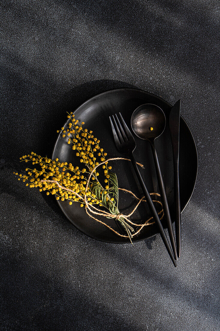 Top view of sophisticated black dining setup featuring a plate, utensils, and a pop of color from bright yellow mimosa flowers against a dark textured background.