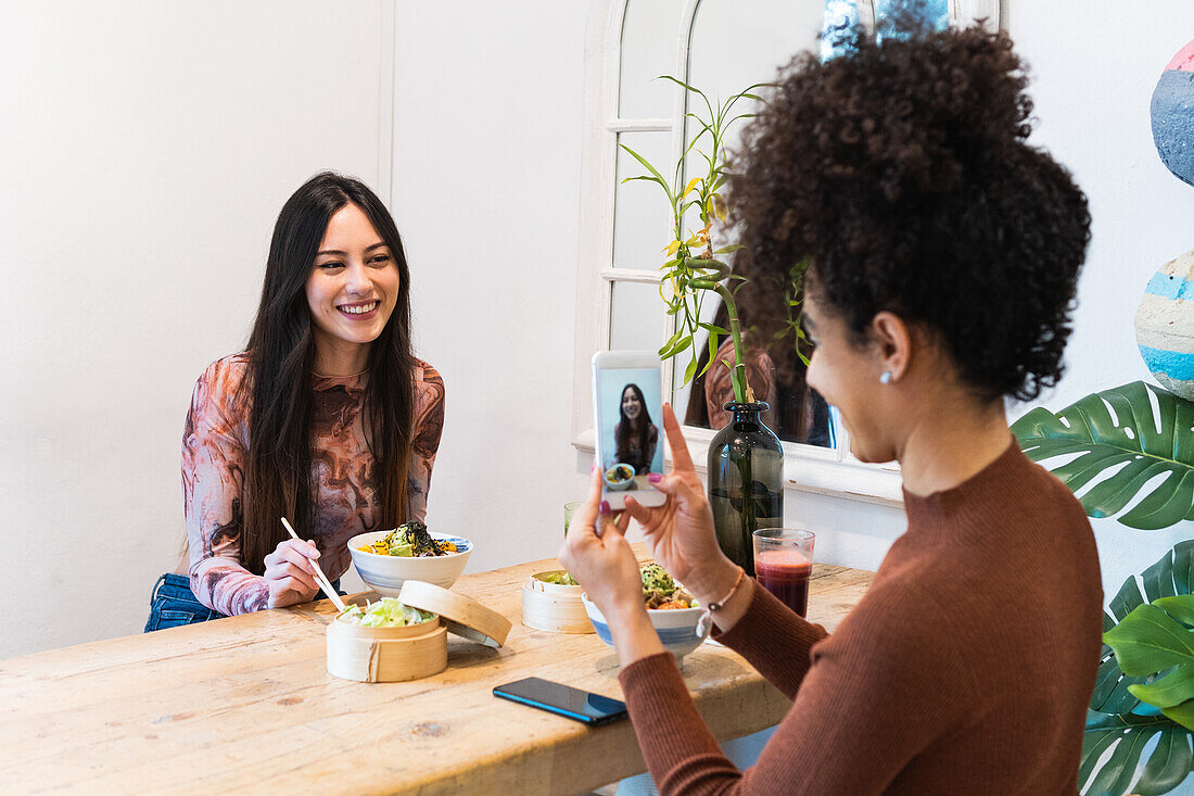 Unrecognizable African American female taking photo of friend sitting at table with poke dish in restaurant