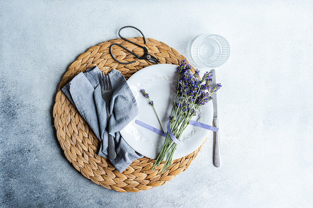 An inviting summer table setting featuring a woven placemat, white dinnerware, linen napkin, cutlery, a glass, and fresh lavender flowers