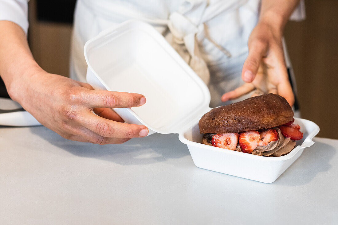 Ein nicht erkennbarer Bäcker, der süßen veganen Biskuit mit Schlagsahne und Erdbeeren in einen Behälter für Speisen zum Mitnehmen in einer Bäckerei legt