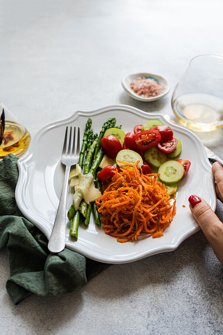 Top view of cropped unrecognizable hand holding a gourmet vegetable plate featuring asparagus with cheese, cherry tomatoes, cucumber slices, and grated carrot, paired with a glass of white wine.