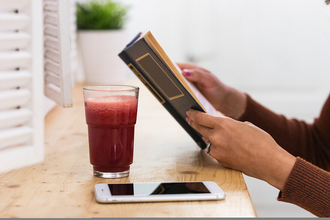 Cropped unrecognizable black female sitting at counter in bar while reading book and drinking refreshing cocktail