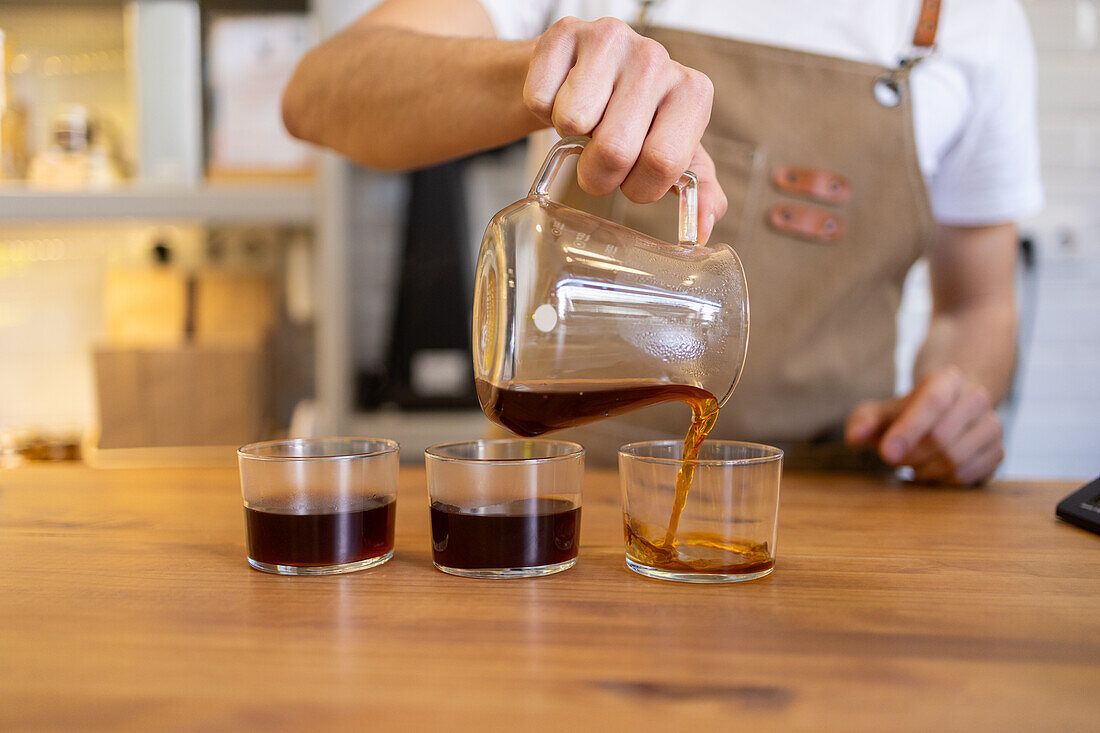 A barista in a bright cafe setting carefully pours steaming coffee into glasses from a clear decanter.