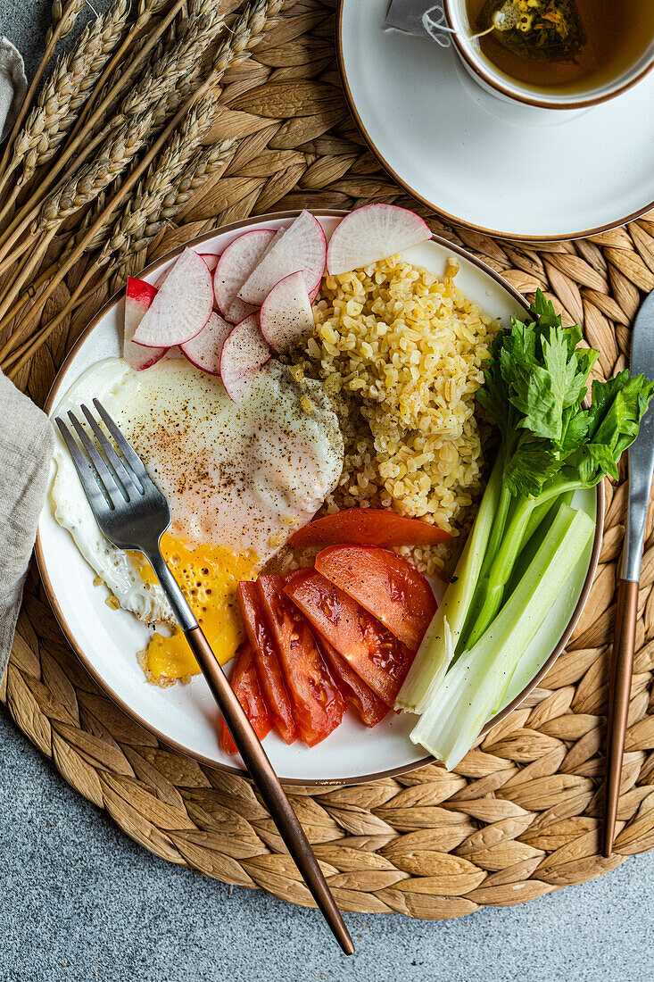 Eine nahrhafte Mahlzeit mit in Scheiben geschnittenen Röteln, Tomaten, Sellerie, einem Spiegelei und Bulgur-Getreide, begleitet von einer Tasse grünem Tee, angerichtet auf einem gewebten Platzdeckchen.