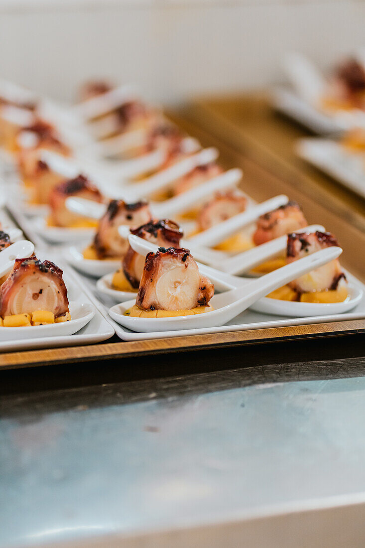 Succulent grilled octopus served over diced fruit, artistically plated on individual white spoons for a sophisticated dining experience