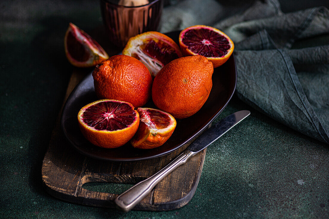 Whole and sliced blood oranges displayed on a dark plate with a vintage knife, against a deep green textured background