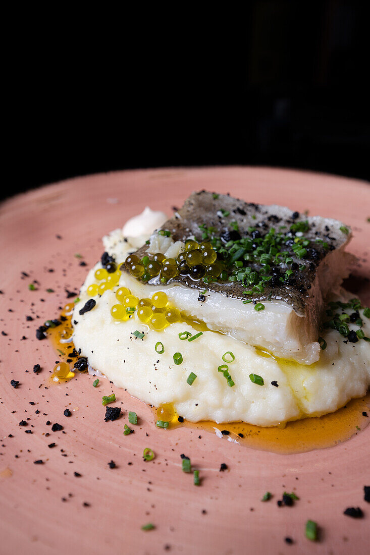 Succulent cod confit laying on a bed of creamy mashed potatoes, garnished with fresh chives, black sesame, and vibrant olive oil pearls, presented on a rustic pink plate.