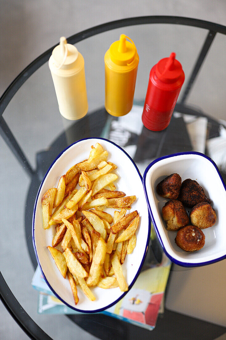 Von oben Genießen Sie einen gemütlichen Tag zu Hause mit einer Portion knuspriger Pommes frites und leckerer Falafelbällchen, die bequem auf einem modernen gläsernen Couchtisch platziert sind, begleitet von Gewürzspendern.