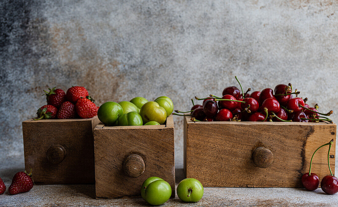 Vintage-Holzkisten mit frischen Bio-Beeren - grüne Pflaumen, süße Kirschen und Erdbeeren vor einem rustikalen Hintergrund, der an den Sommer erinnert