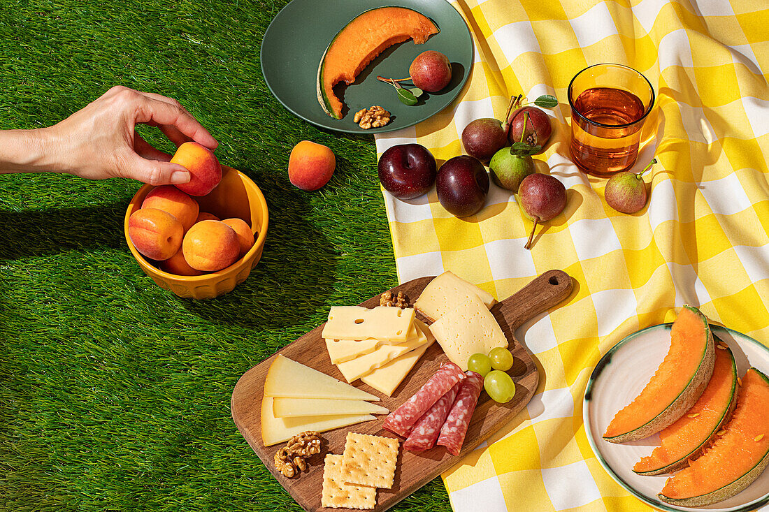 Anonymous person's hand is selecting an apricot from a basket at an outdoor picnic featuring an assortment of cheese, meats, fruits, and a drink on a checkerboard blanket