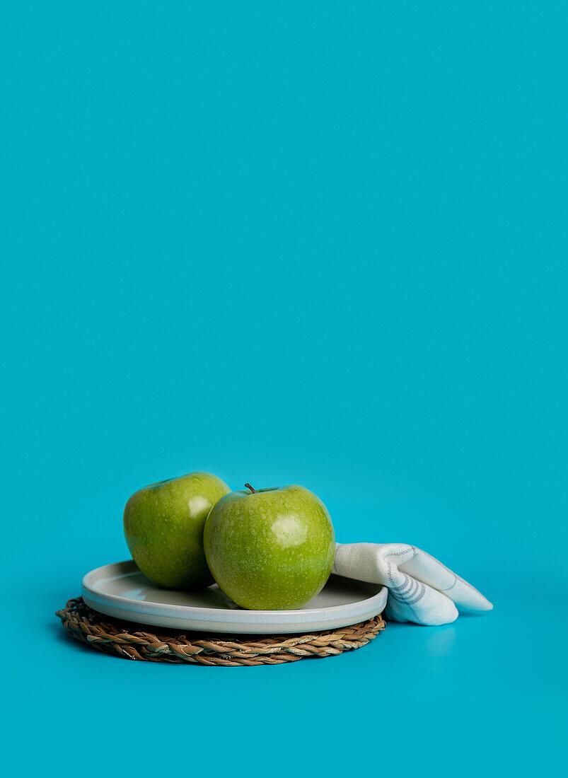 Two vibrant green apples neatly placed on a gray plate over a woven round mat. This composition showcases a simplistic yet appealing approach to presenting healthy and wholesome food, emphasized by a striking blue background.