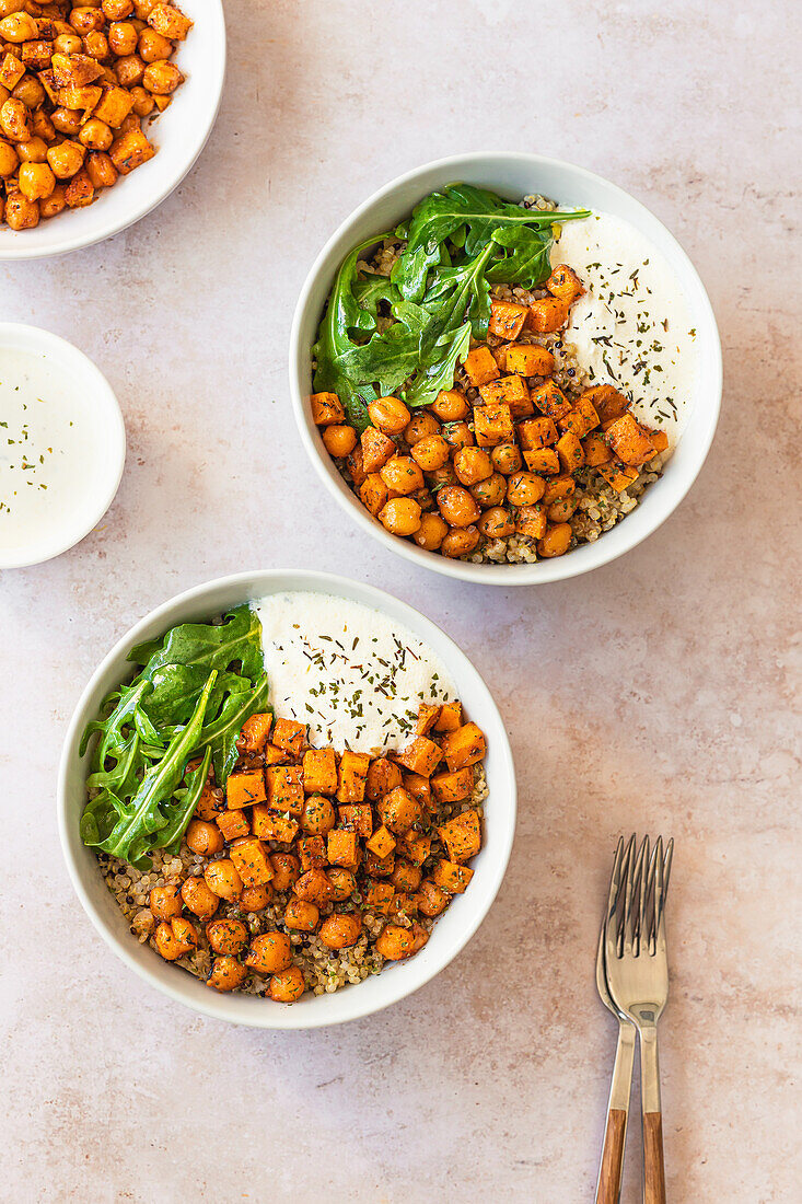 Two bowls of nutritious quinoa salad topped with spiced chickpeas, roasted sweet potato cubes, fresh arugula, and a dollop of creamy yogurt sauce, garnished with dried herbs