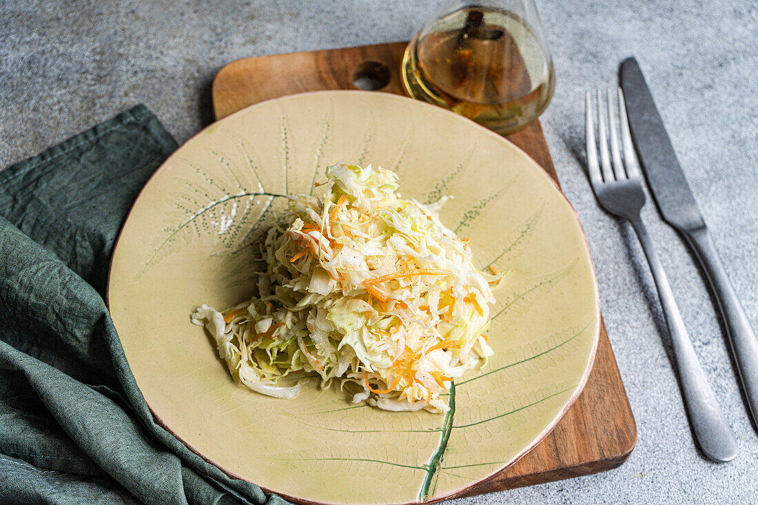 Top view of a healthy organic cabbage and carrot salad drizzled with olive oil, served on a decorative ceramic plate, accompanied by a glass bottle of olive oil and utensils on a textured surface.