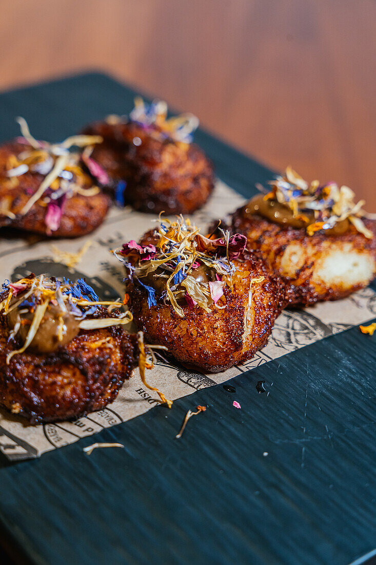 Exquisite crispy patties are garnished with edible flowers and sauce, presented on a slate board for a refined culinary display