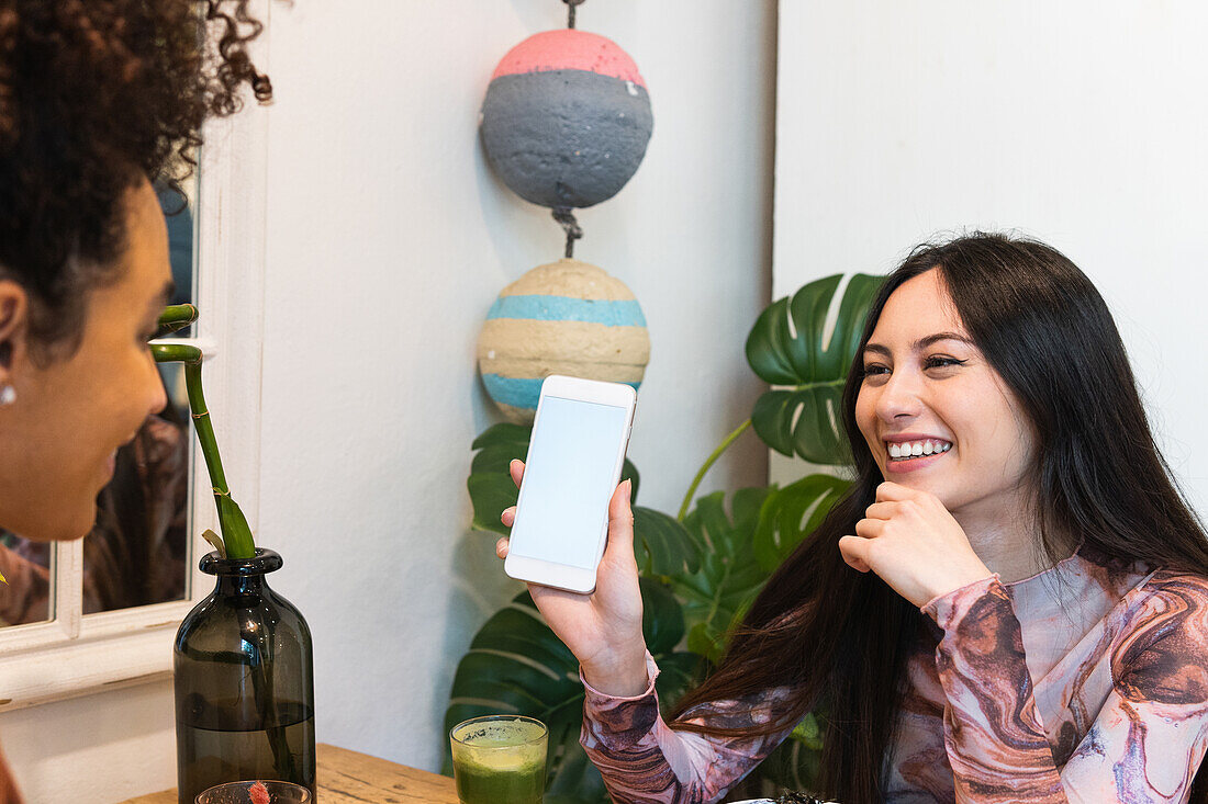 Cheerful diverse female friends sitting at table with poke in restaurant and using smartphone while spending weekend together