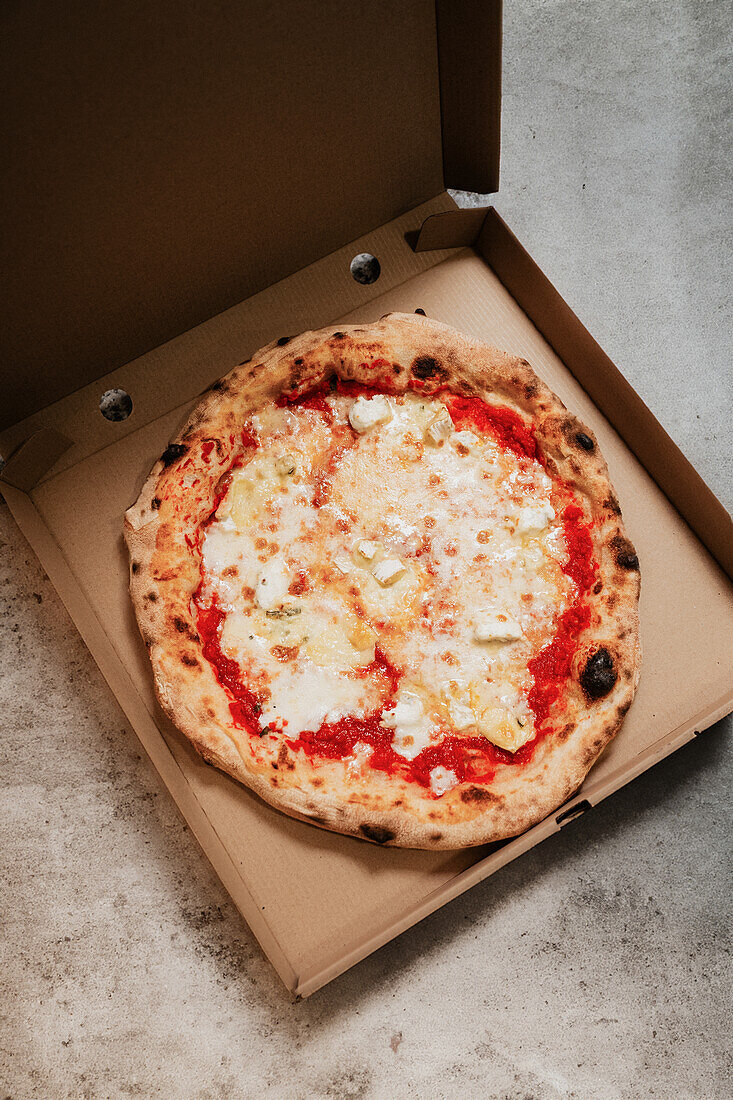 An overhead view of a freshly baked pizza inside a cardboard box, placed on a textured concrete surface, representing takeaway or delivery from a pizzeria