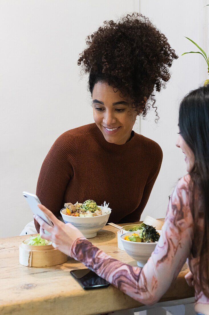 Cheerful ethnic woman sitting at table with crop anonymous female friend eating poke in restaurant and using smartphone while spending weekend together