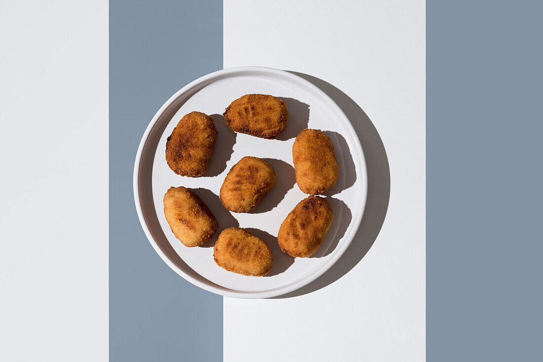 Artistic overhead shot of golden fried croquettes arranged on a white circular plate, half-covered by a long shadow