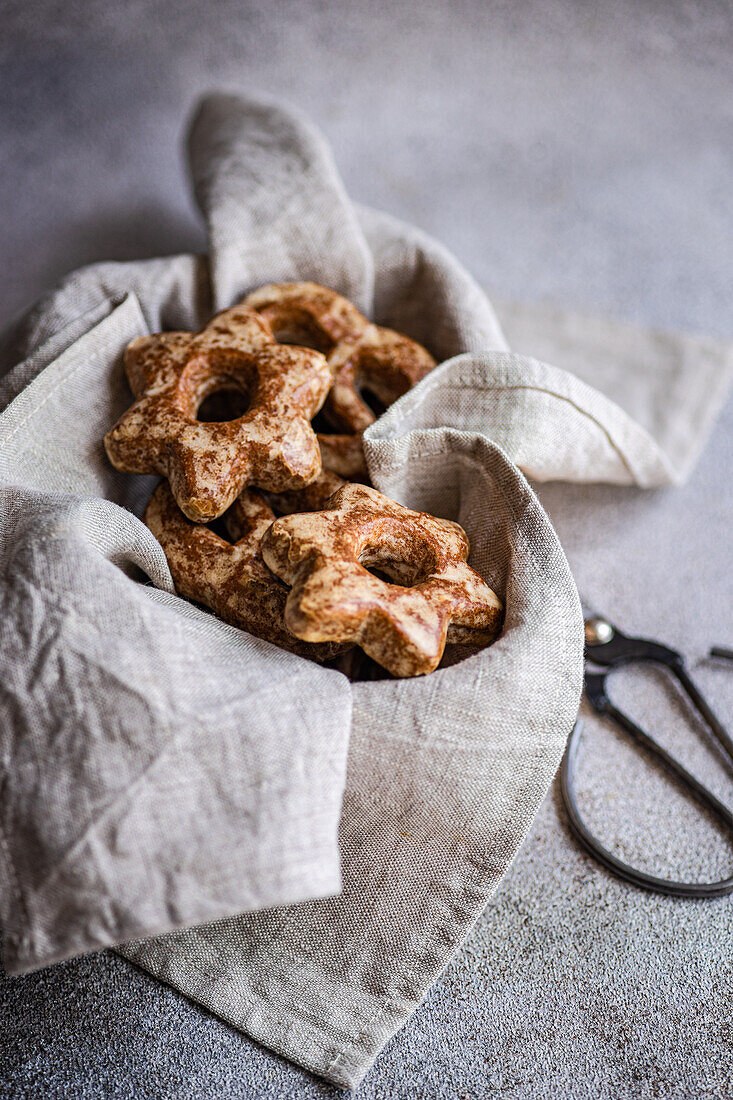 Blick von oben auf selbstgebackene Lebkuchen in Sternform, die in ein graues Tuch eingewickelt sind und eine gemütliche und rustikale Atmosphäre schaffen. Eine Schere verleiht der Szene einen Hauch von Handwerk und unterstreicht die handwerkliche Qualität.