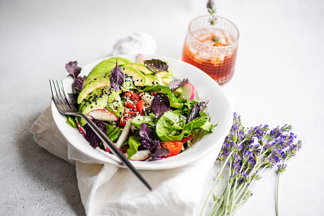 A colorful fresh vegetable salad featuring lettuce, olives, cherry tomatoes, cucumbers, radishes, and red basil, sprinkled with white and black sesame seeds, presented in a white bowl with a fork.